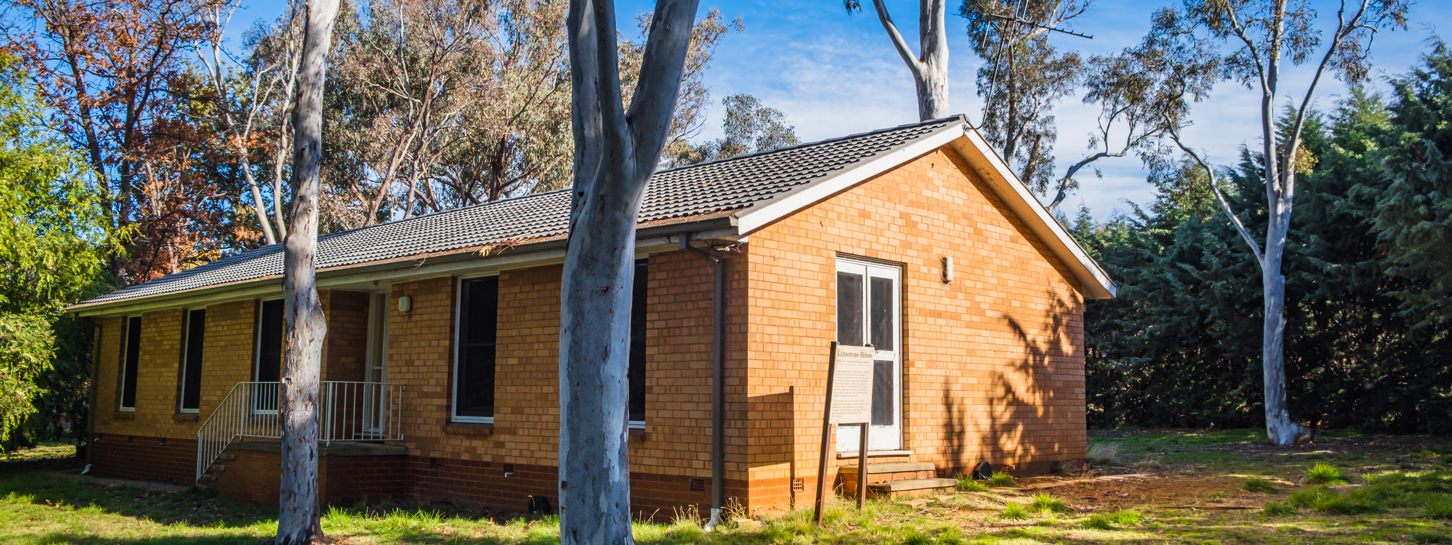 Photo of an orange brick house, with a brown tiled roof and white edged glass windows, surrounded by grass and gumtrees, with blue sky in the background.