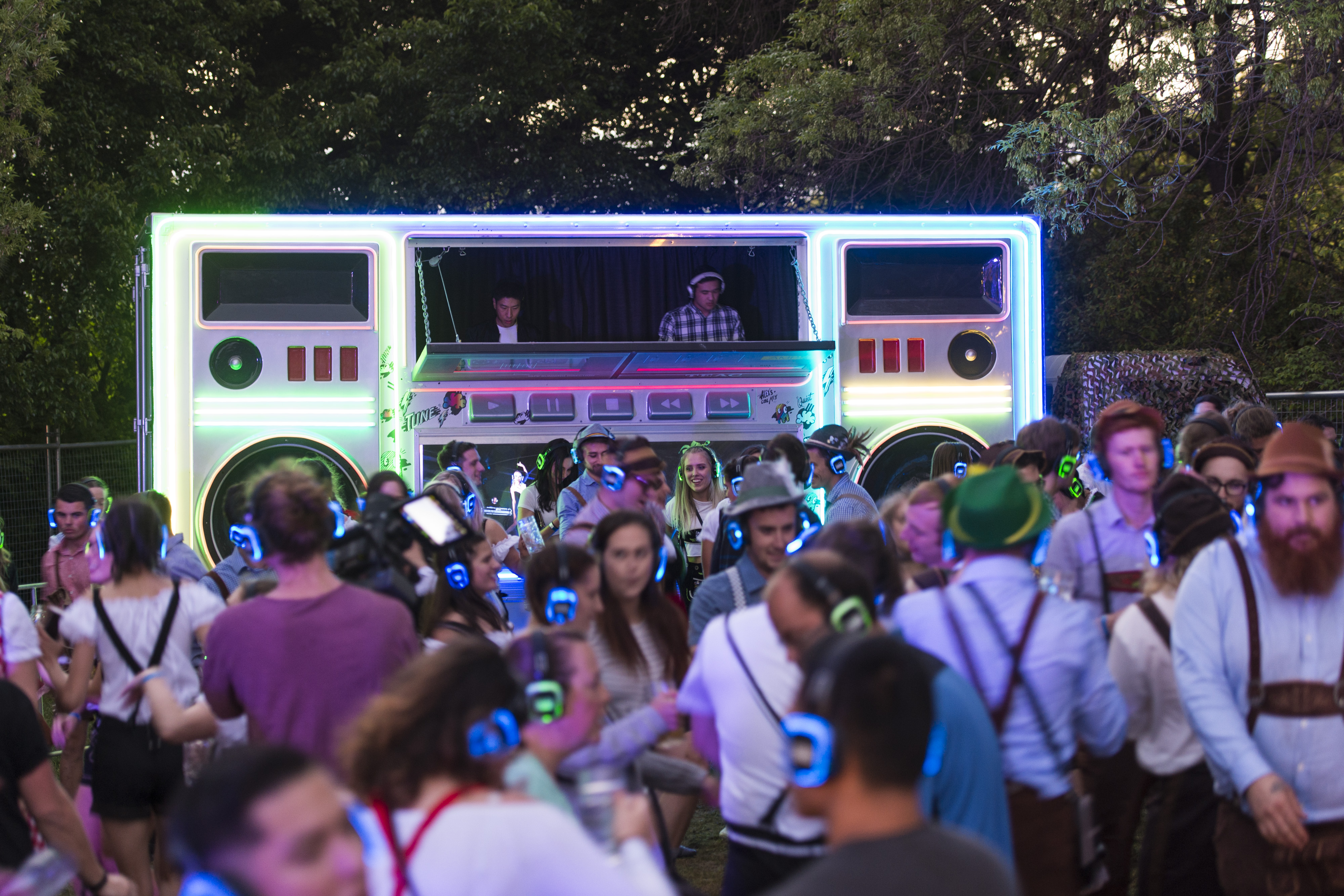 Festival attendees wearing illuminated wireless headphones gather around a neon-lit DJ booth designed to resemble a giant retro cassette player. The outdoor evening scene is set among trees, with people socialising and dancing during a silent disco event.