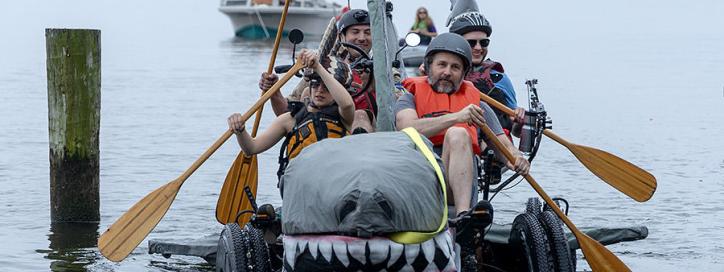 Two men and two women in life vests rowing a giant shark on wheels through water