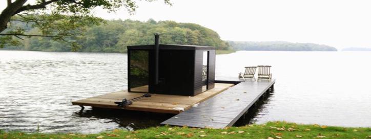 photo of black exterior of a sauna floating on a wooden pontoon on the lake edging the grass with a tree framing the image with lush green leaves and a curve of blurred land mass in the far distance
