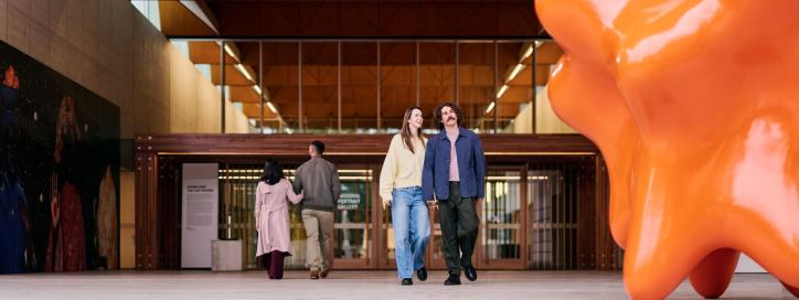 Modern gallery entrance with visitors walking past a large orange sculpture and contemporary artwork.