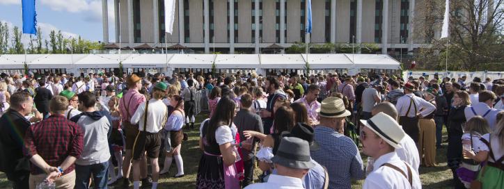 Large crowd attending an outdoor festival in front of a modern civic building under a bright blue sky. Long white vendor tents line the event area while attendees gather across the lawn, many dressed in traditional Oktoberfest-style attire.