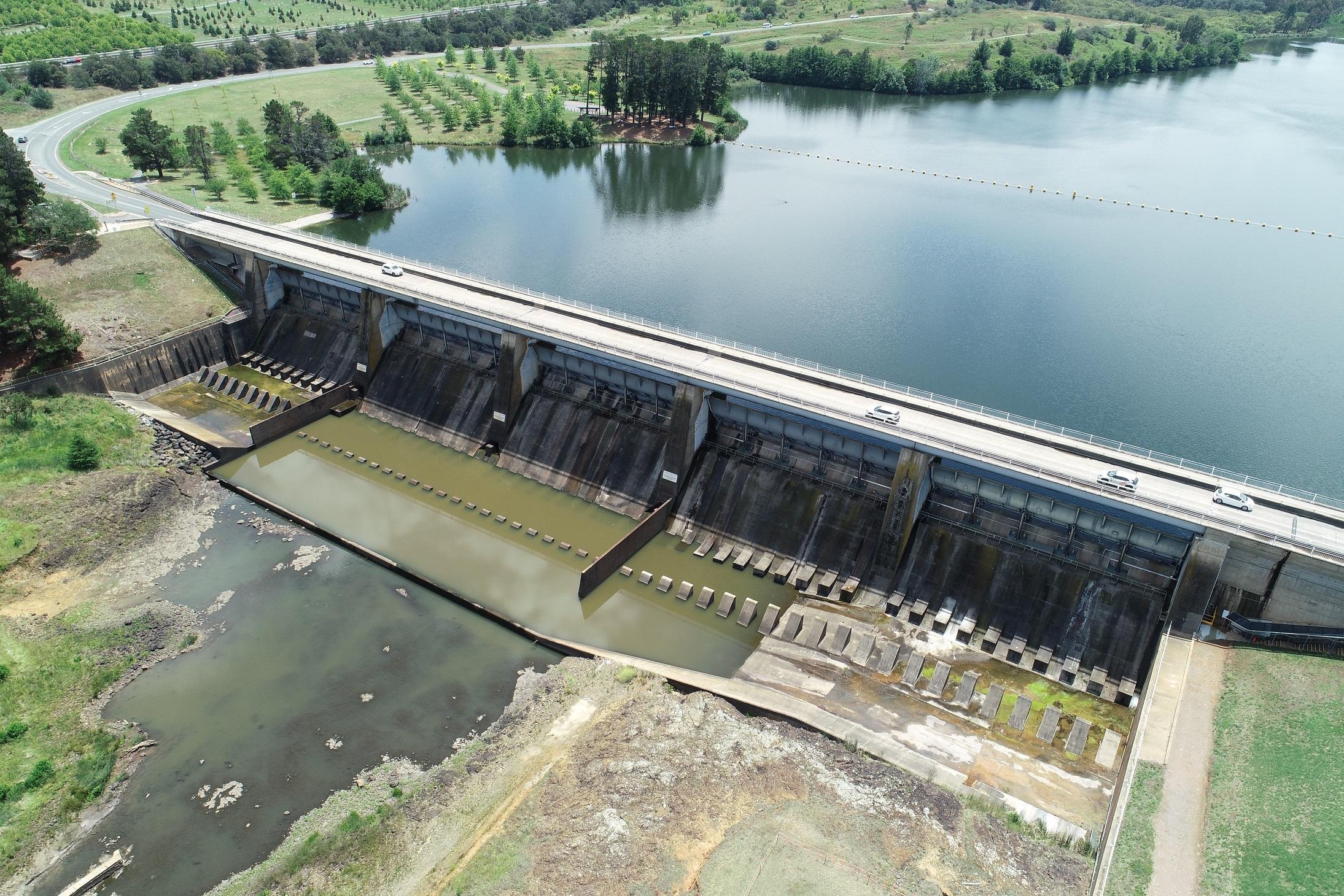 This image is a drone photo of Scrivener Dam from above showing the road bridge and water in the base of the dam with Lake Burley Griffin in the background.
