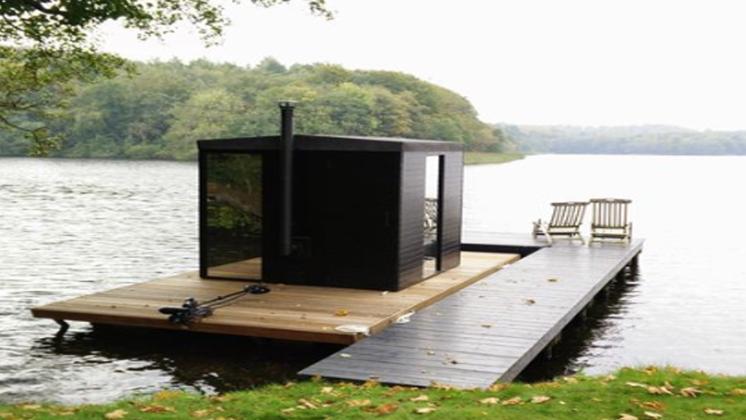 photo of black exterior of a sauna floating on a wooden pontoon on the lake edging the grass with a tree framing the image with lush green leaves and a curve of blurred land mass in the far distance