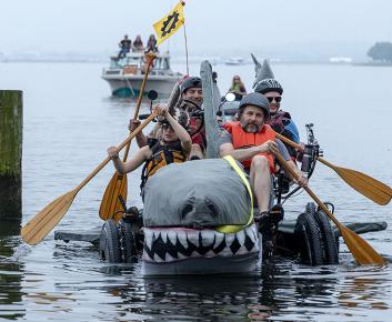 Two men and two women in life vests rowing a giant shark on wheels through water