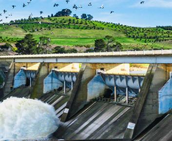 Photo of Scrivener Dam on a blue sunny day with water flowing through at high pressure creating a plume of mist and water at the bottom of the dam release