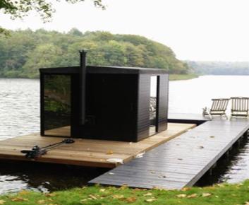 photo of black exterior of a sauna floating on a wooden pontoon on the lake edging the grass with a tree framing the image with lush green leaves and a curve of blurred land mass in the far distance