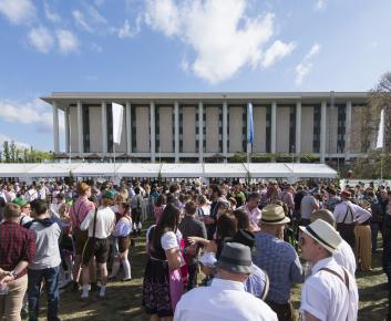 Large crowd attending an outdoor festival in front of a modern civic building under a bright blue sky. Long white vendor tents line the event area while attendees gather across the lawn, many dressed in traditional Oktoberfest-style attire.